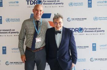 Dr. Diemert and colleague smiling in front of the TDR step and repeat banner 